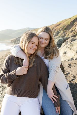 Two friends share an affectionate moment while sitting together on a rocky beach at sunset.