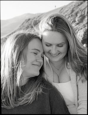 Black and white portrait of two women smiling and leaning against each other with windswept hair outdoors.