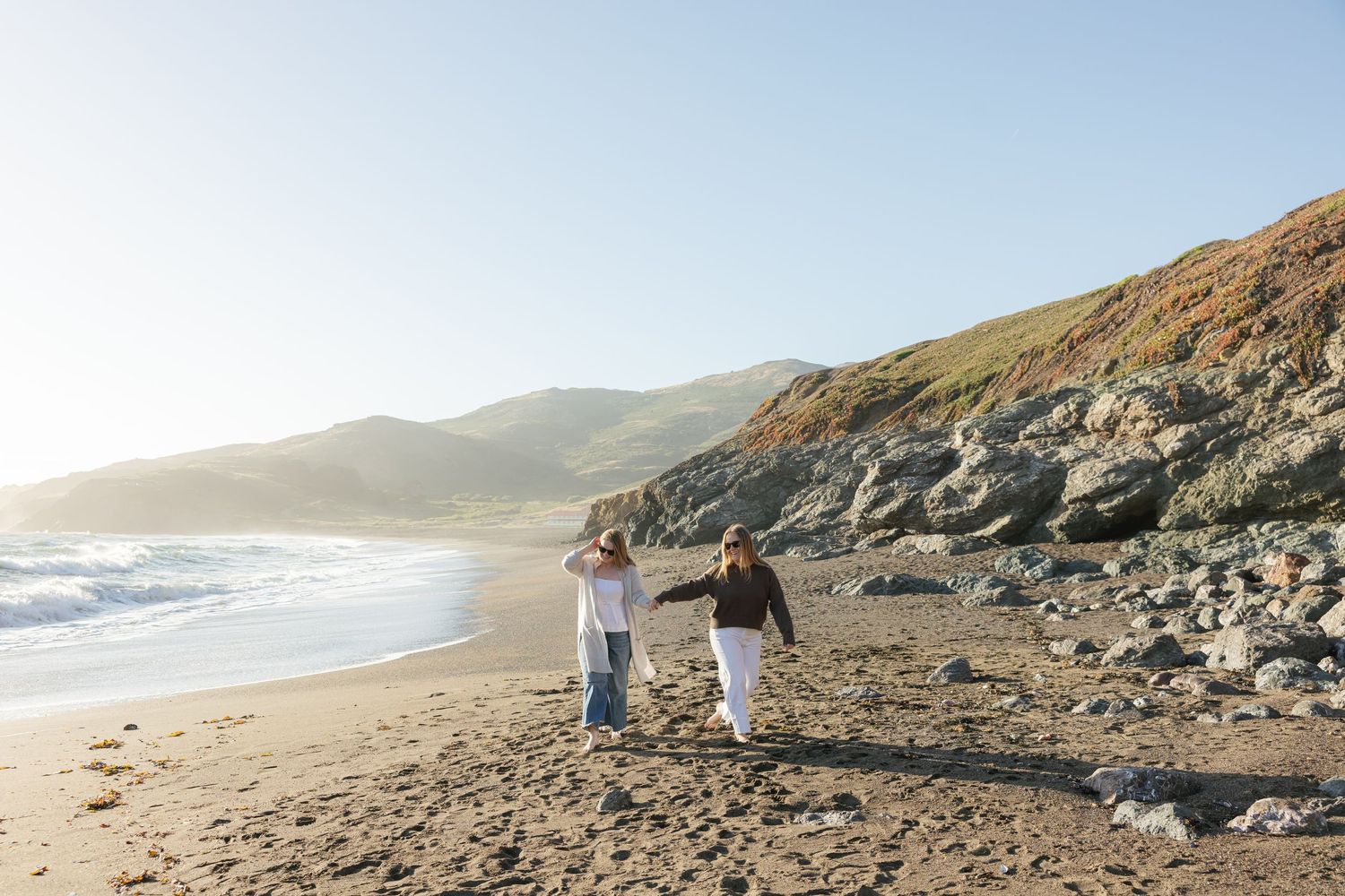 Two hikers walk along rugged Pacific coastline with mountains and rocky cliffs visible in golden afternoon light.