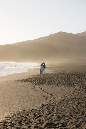 Silhouetted couple walking on sandy beach at sunset with mountains in background.