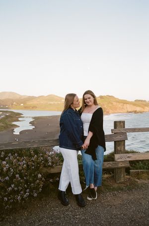 Two friends lean against a wooden railing overlooking a scenic coastal landscape at sunset.