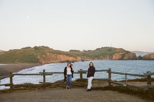 Two people stand at a coastal viewpoint overlooking dramatic cliffs and ocean waves during golden hour.