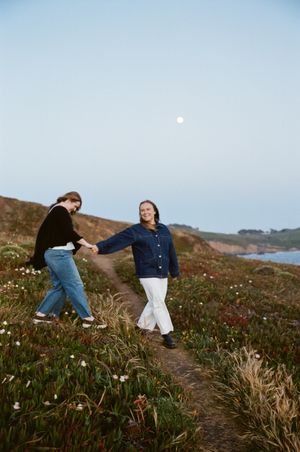 Two people share a playful moment on a coastal hillside with wildflowers at sunset.