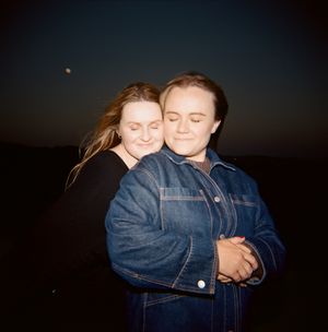 Two people embrace warmly while wearing denim jackets at dusk with a dark sky backdrop.