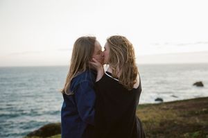 Two people share an intimate moment on a coastal cliff overlooking the ocean at sunset.