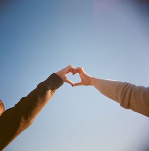Two hands reach up against a blue sky to form a heart shape together in warm sunlight.