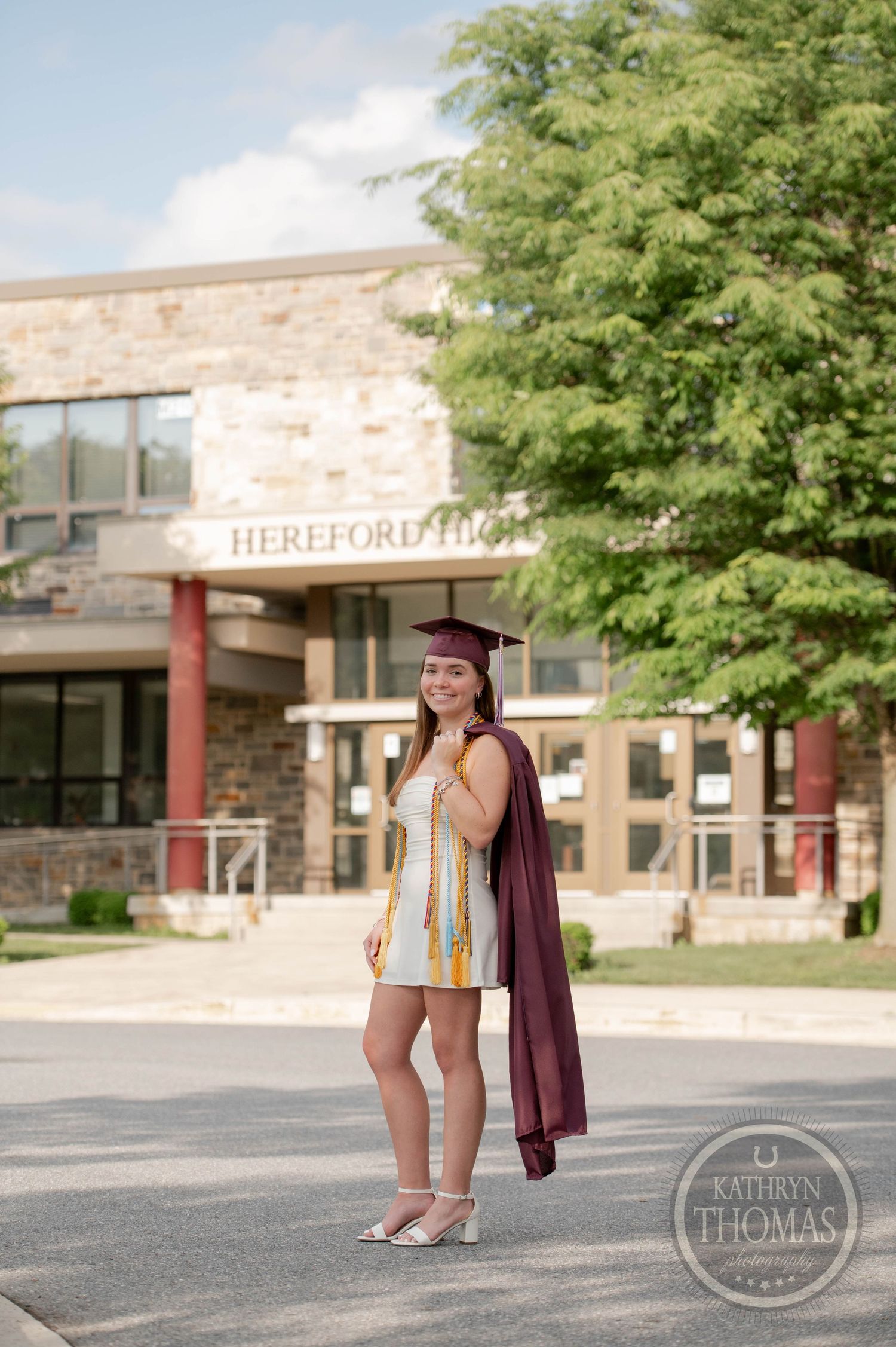 Graduate in burgundy cap and gown stands outside Hereford Hall on college campus in summer.