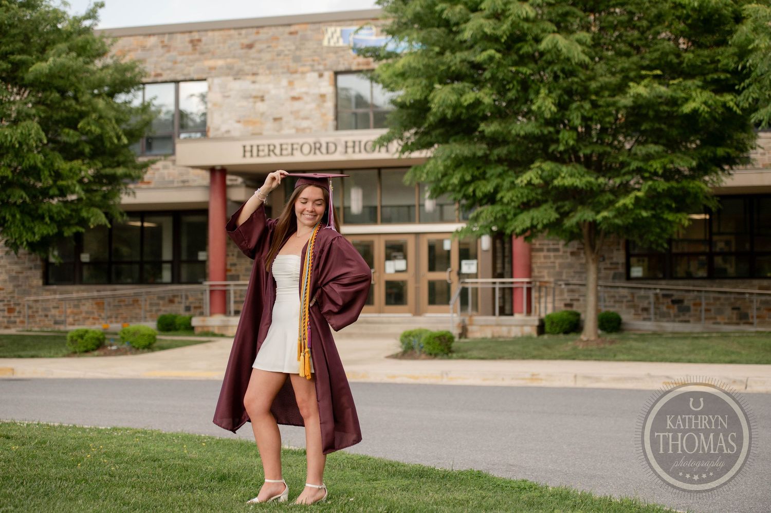 A person wearing a burgundy coat stands outside a stone building entrance with mature trees on a sunny day.