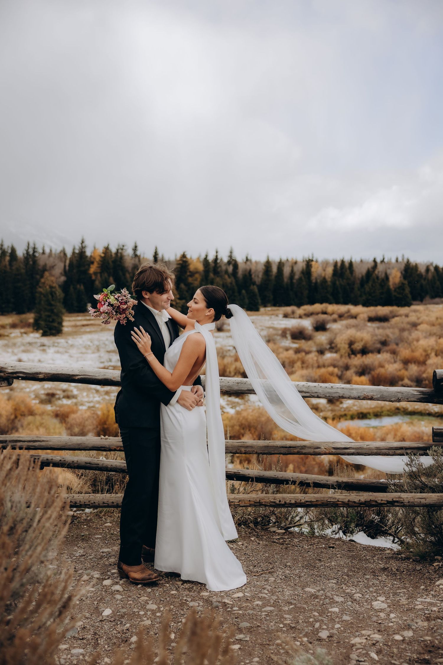 A romantic wedding couple embraces against an autumn forest backdrop with flowing white veil in the mountain breeze.