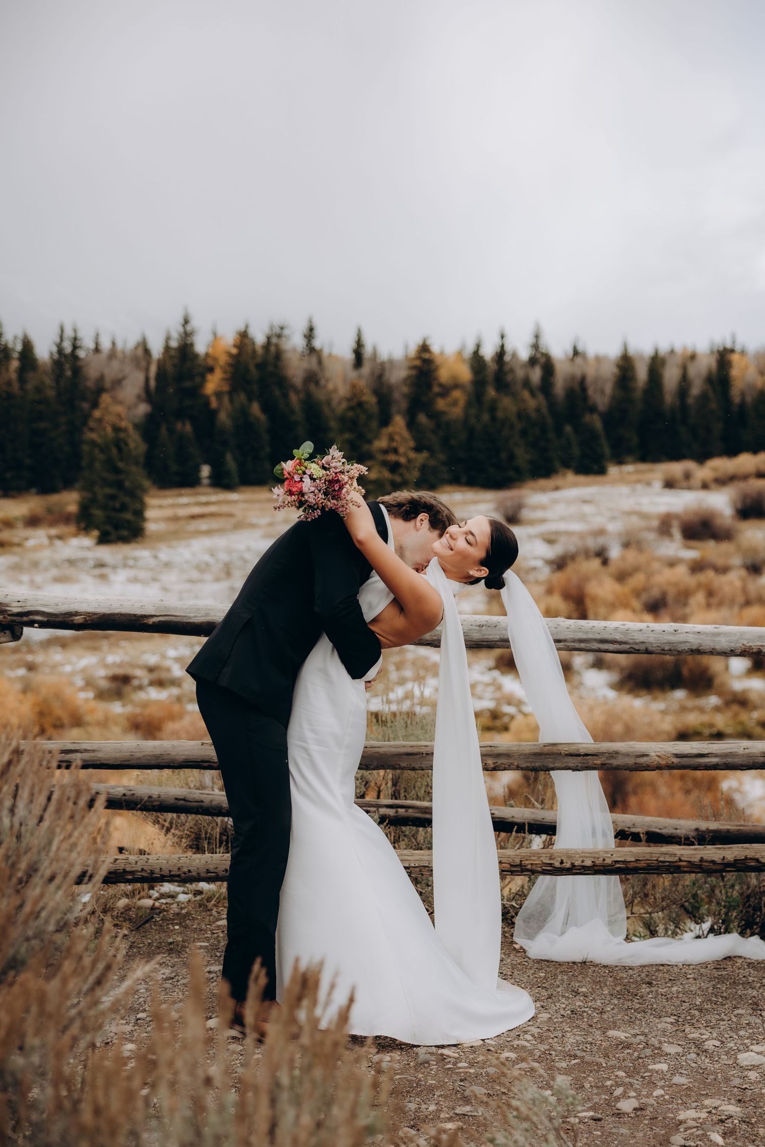 Romantic wedding couple dips and dances against a backdrop of evergreen trees and rustic wooden fencing at sunset.