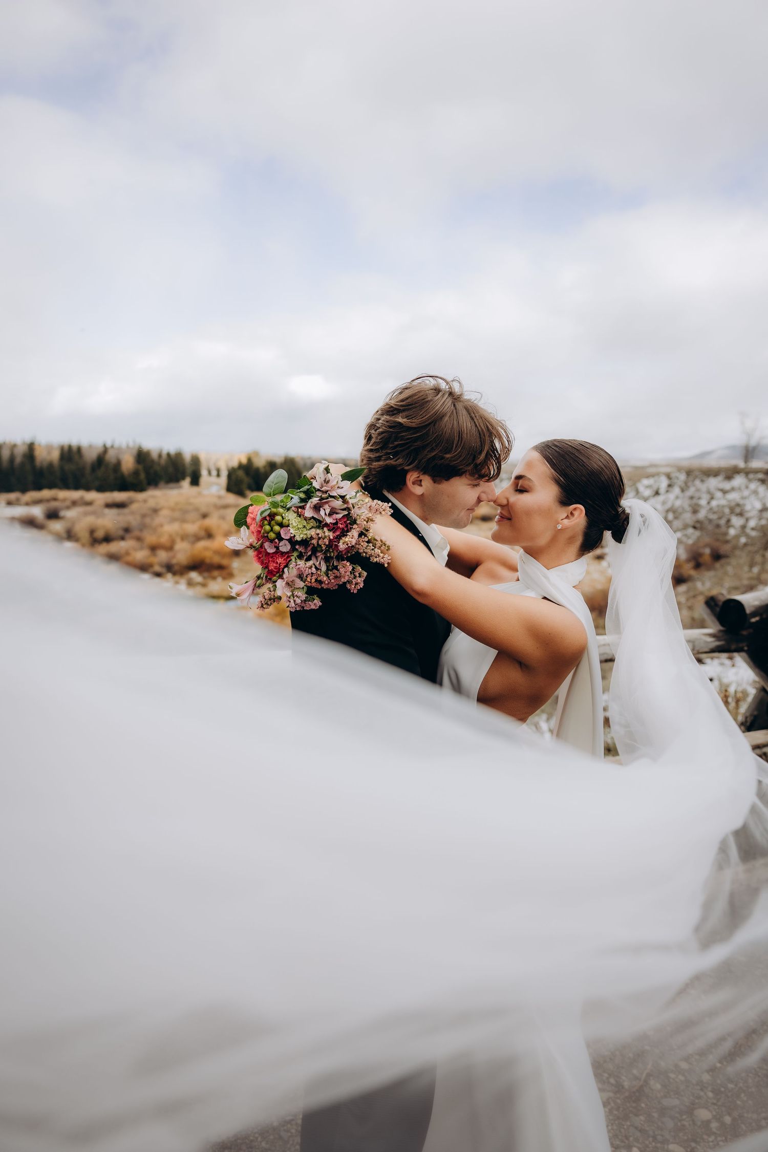 A romantic wedding couple embraces outdoors with a flowing white veil and wildflower bouquet against a scenic landscape.