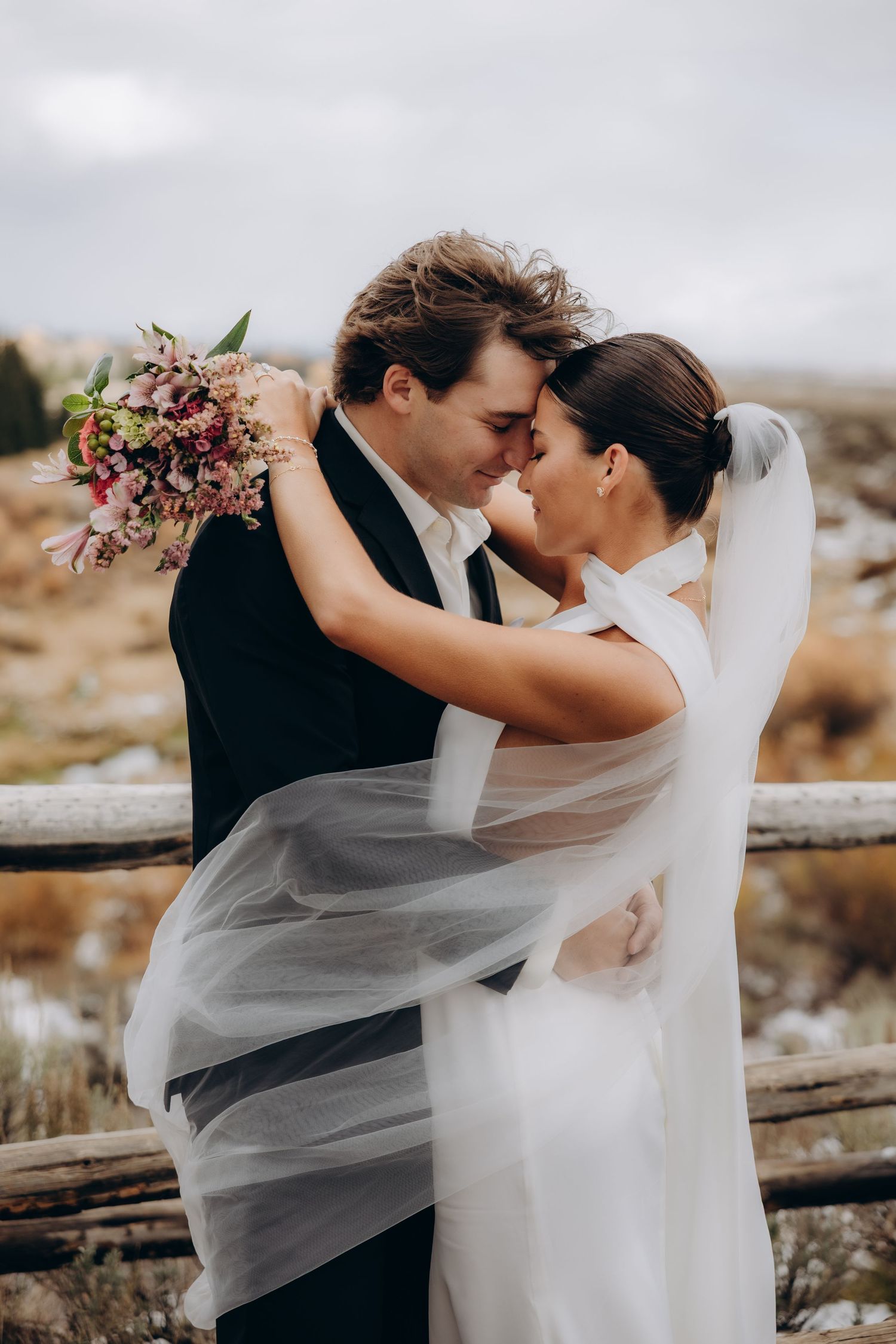 A romantic wedding couple embraces outdoors in a rustic setting with a flowing veil and autumn bouquet.