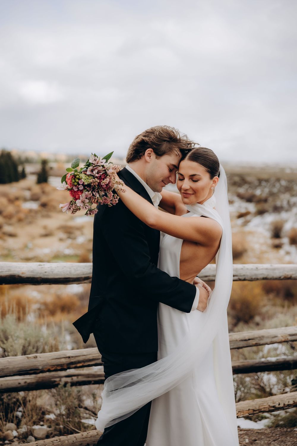 A couple shares an intimate embrace on their wedding day against a rustic wooden fence in a desert landscape.