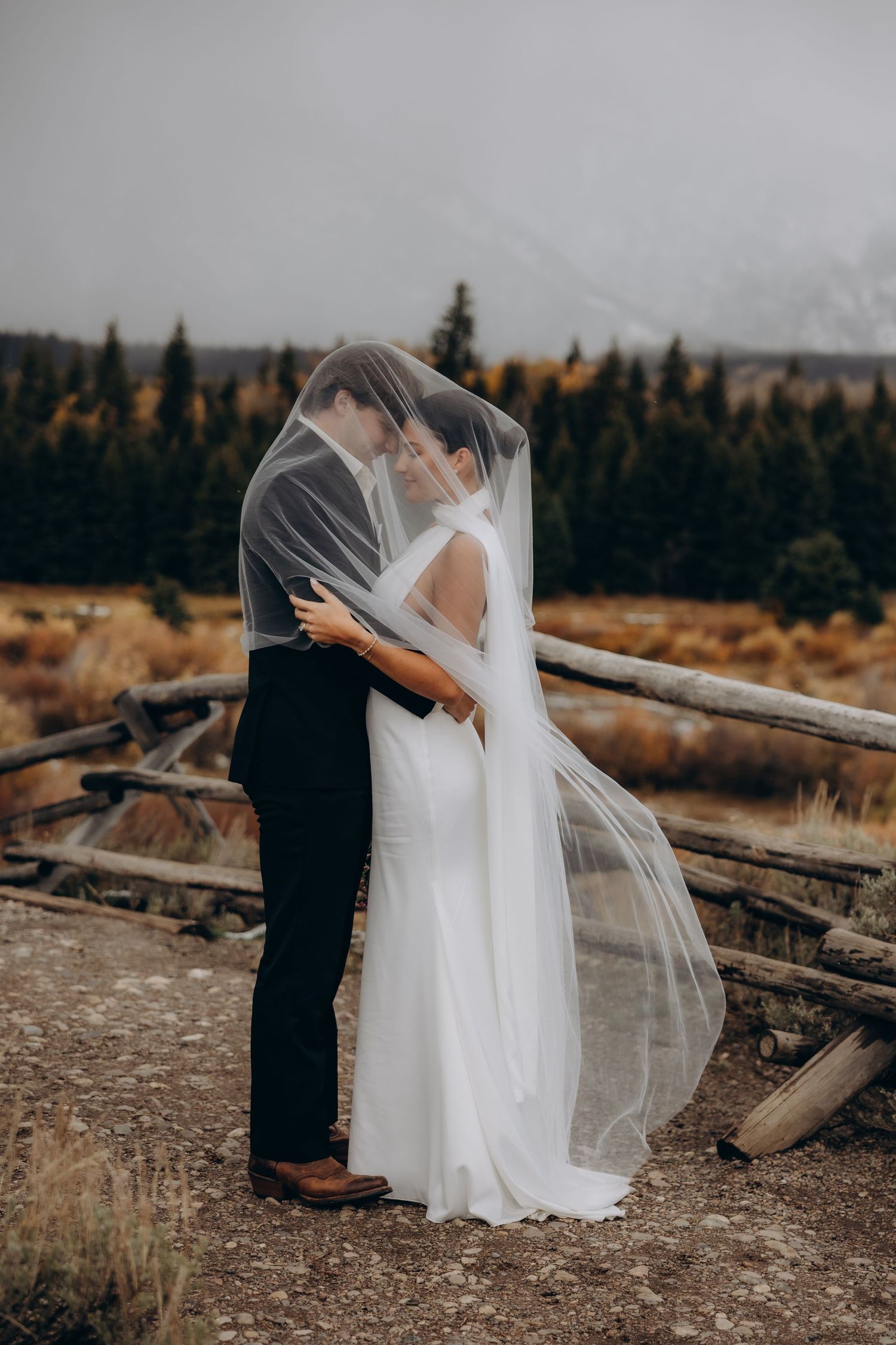 A romantic wedding moment captured on a rugged terrain with driftwood and evergreen forest in the background.