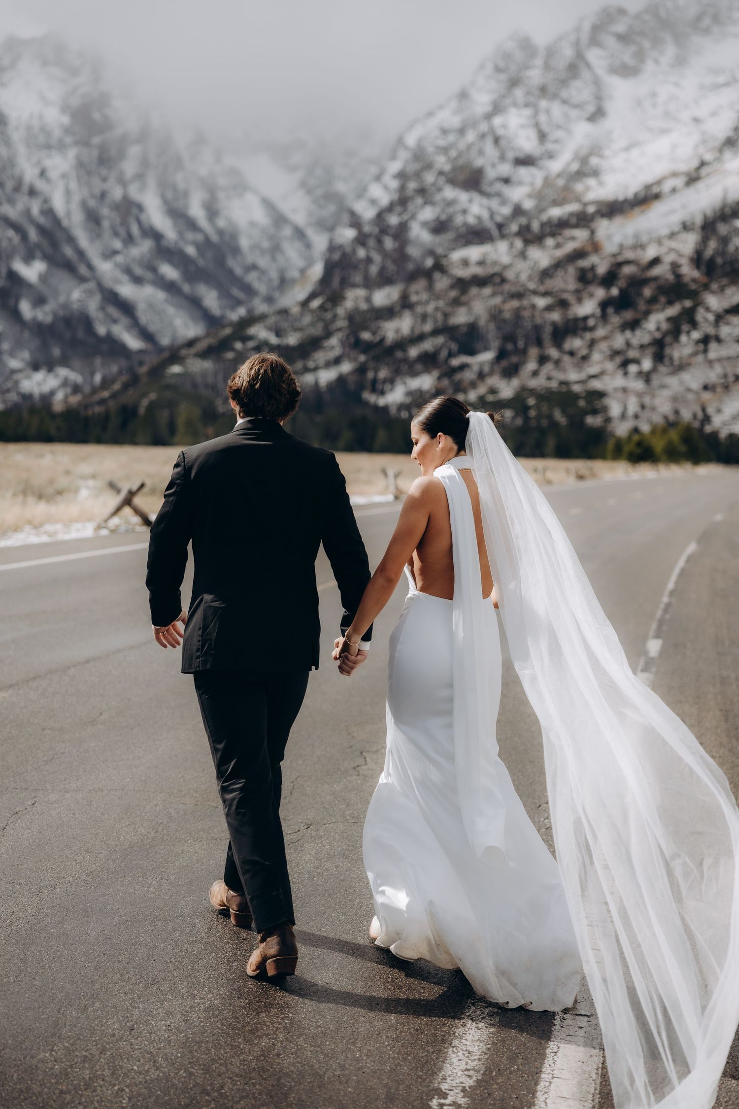 A couple in wedding attire walks hand in hand down a mountain road with snow-capped peaks in the background.