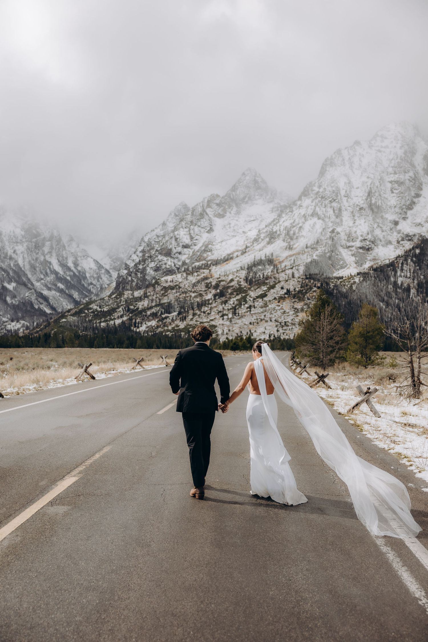 A couple walks along a snowy mountain road with dramatic snow-capped peaks in the background.