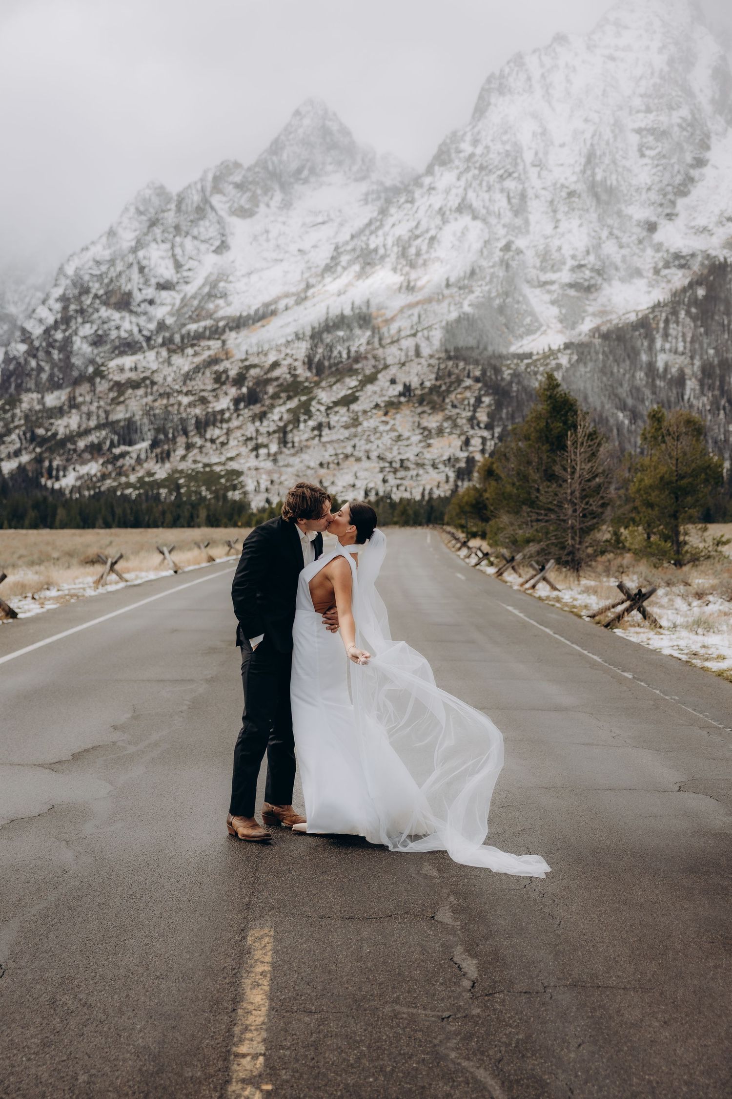 Newlyweds share a romantic moment on an empty mountain road surrounded by majestic snowy peaks.