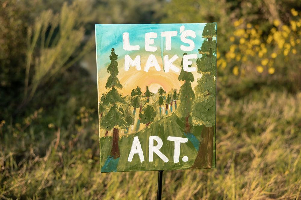 Handmade signs reading 'LETS MAKE ART' displayed in a natural outdoor setting with sunlit foliage in the background.