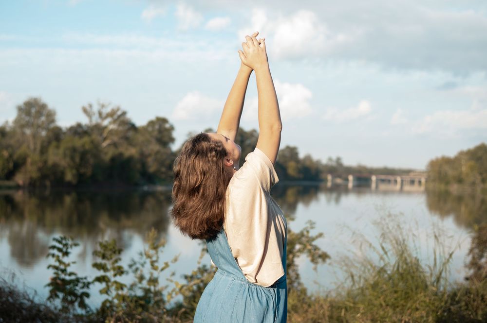 Person stands with raised arms by a serene lake surrounded by trees on a cloudy day.