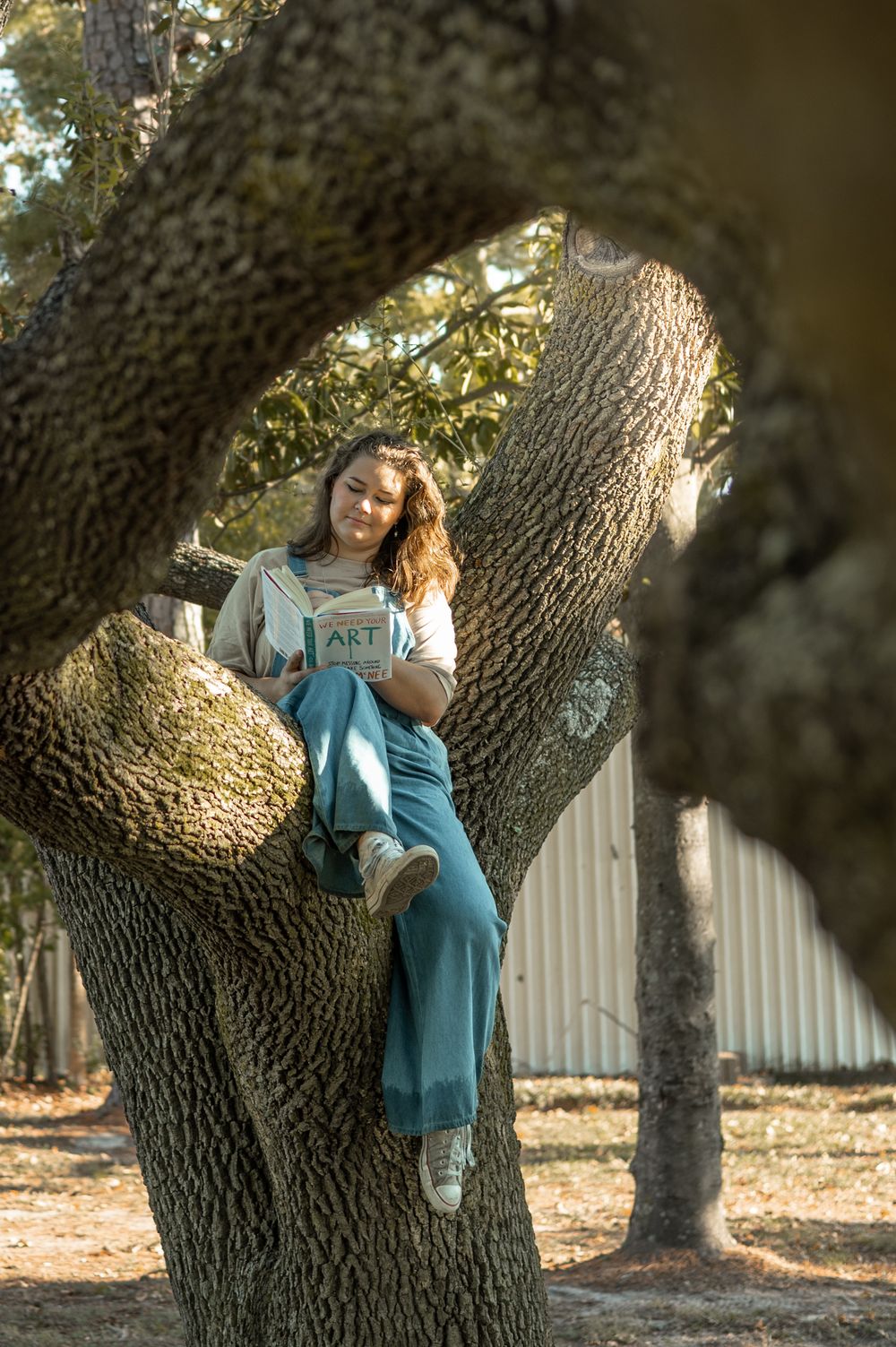 A person in a blue sweater poses thoughtfully against a large tree trunk in dappled sunlight.