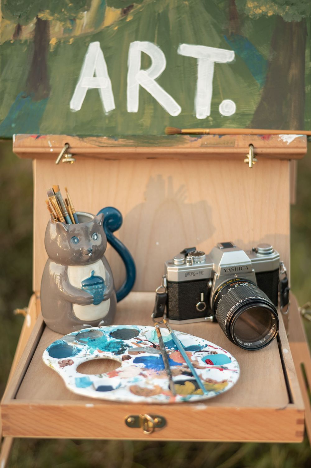 Vintage camera and art supplies arranged on a wooden shelf with blue paint palette and ceramic mug.