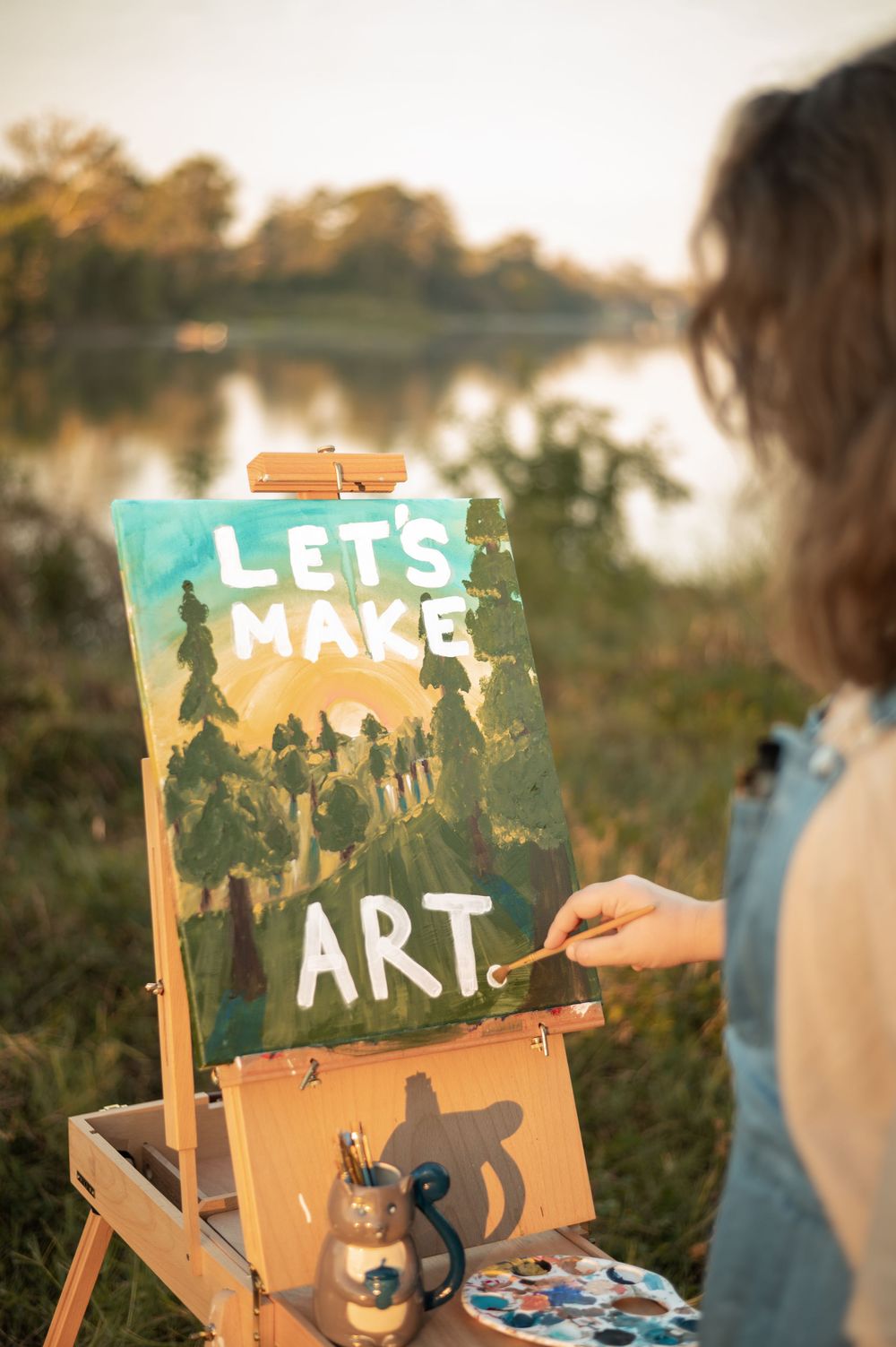 A wooden easel with 'Let's Make Art' text sits in a sunny outdoor setting with trees in the background.