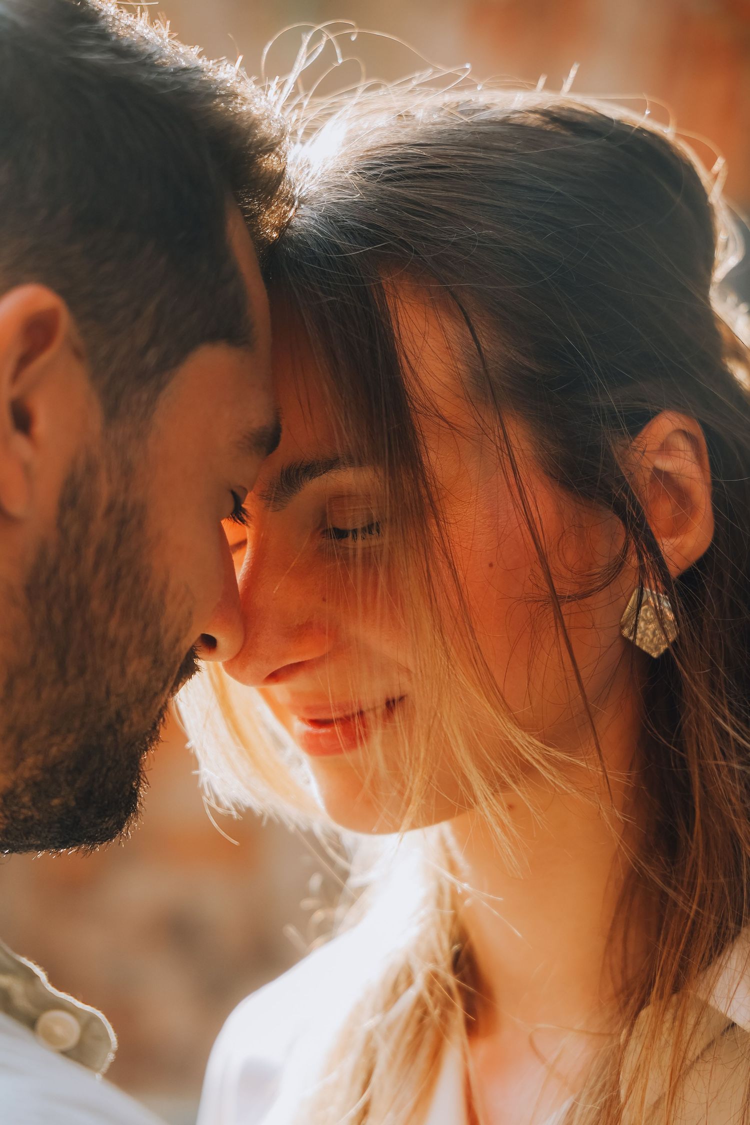 Intimate close-up of couple touching foreheads in golden sunlight, woman wearing decorative earring smiling softly.