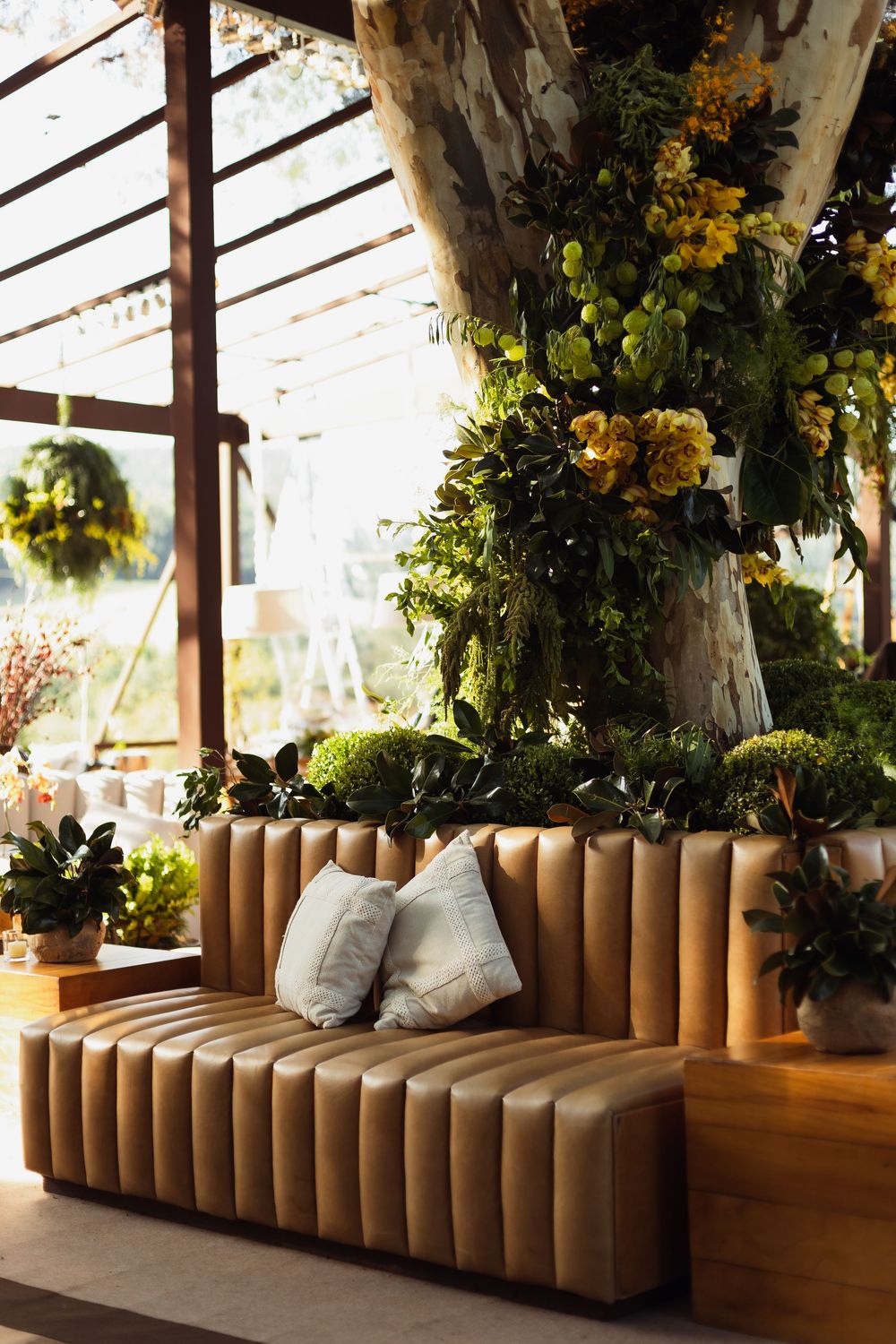 Modern brown velvet sofa with pillows in a greenhouse setting adorned with hanging floral arrangements and greenery.