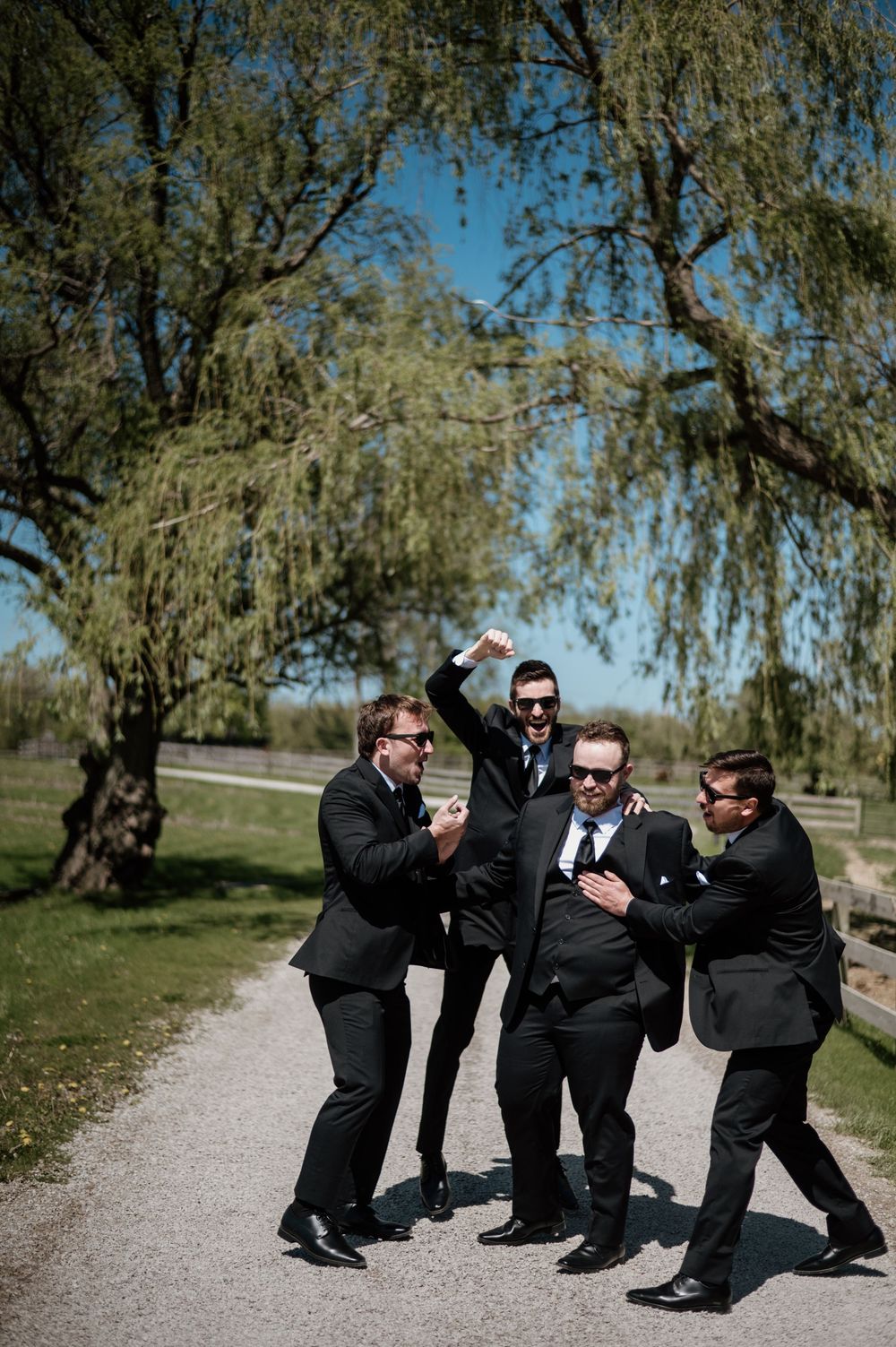 Groomsmen in black suits playfully pose in action movie style on a path surrounded by willow trees.