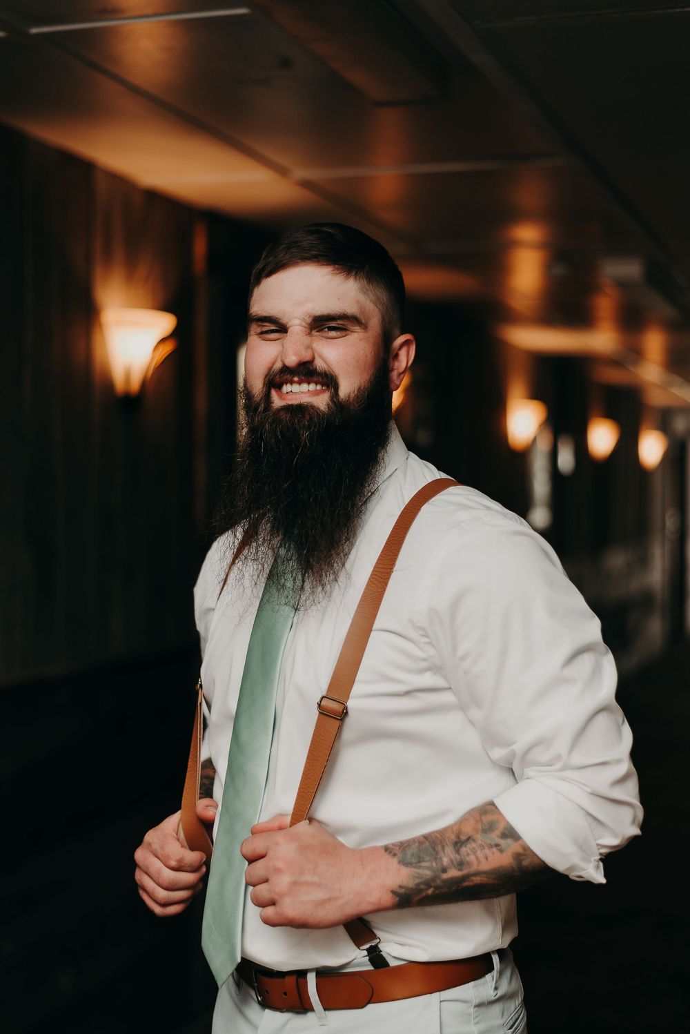 Groom in white shirt and suspenders poses in moody indoor lighting.