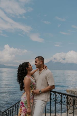 A romantic couple shares tender moments by an oceanside railing under a bright blue sky.
