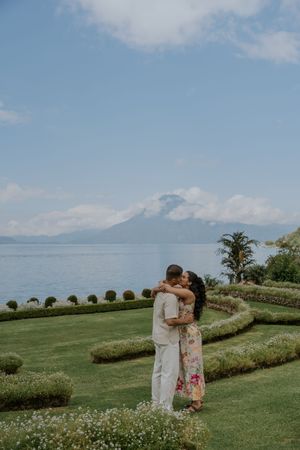 A couple embraces on manicured lawn overlooking Lake Atitlan and volcanic mountains in Guatemala on a sunny day.