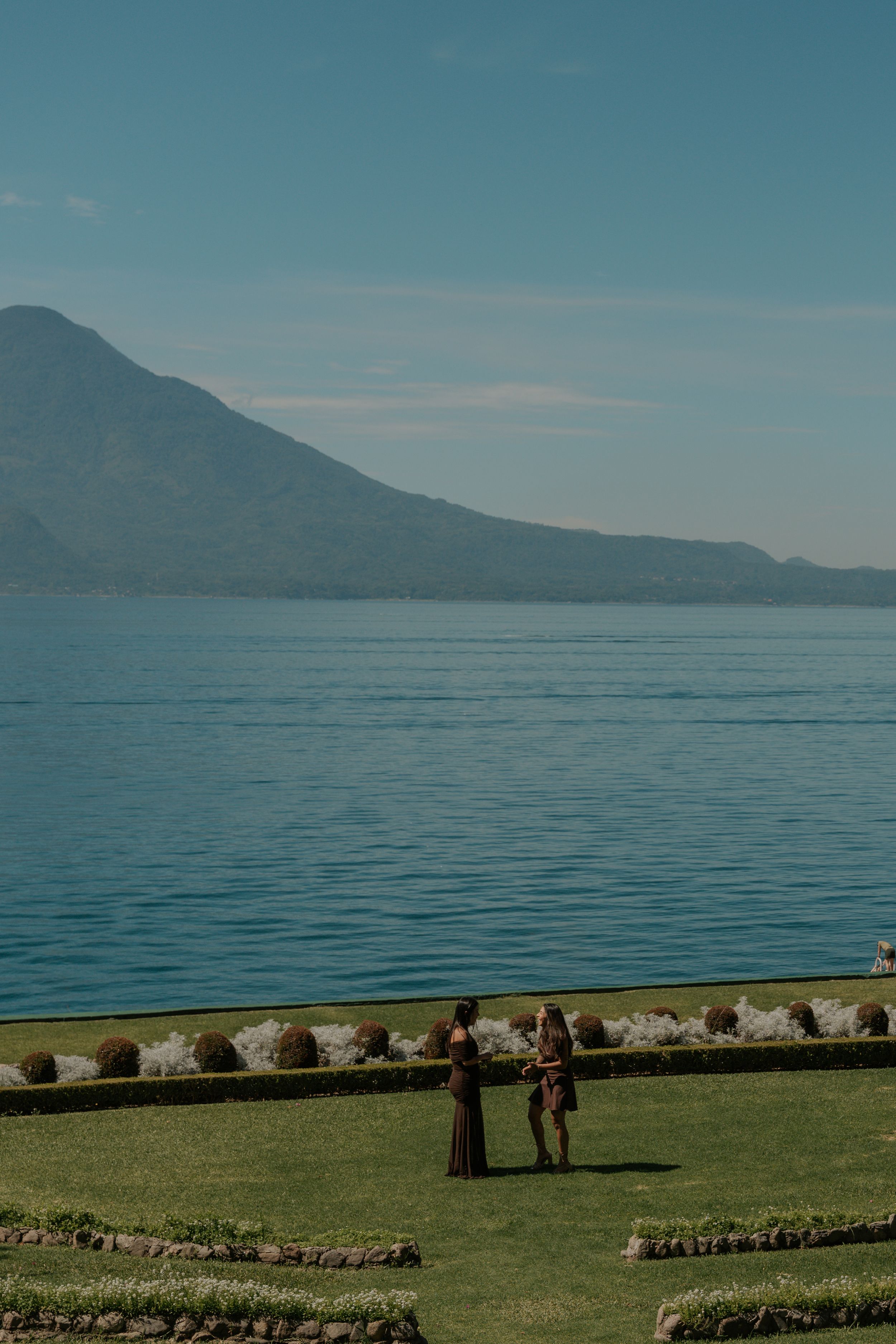 Scenic lakeside view with mountains in the background, featuring a grassy shoreline and stone border under blue skies.