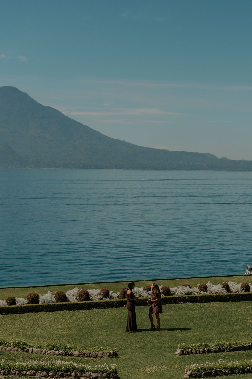 Scenic lakeside view with mountains in the background, featuring a grassy shoreline and stone border under blue skies.