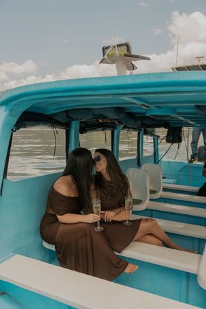 Two friends in brown dresses share champagne inside a turquoise boat on a sunny day.