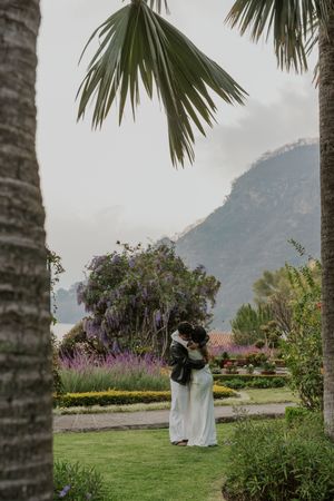 Couple embracing in a lush garden with palm trees, colorful flowers, and misty mountain backdrop.