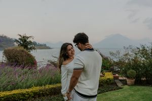 Couple in white outfits share a romantic moment in a lush garden with mountain lake views in the background.
