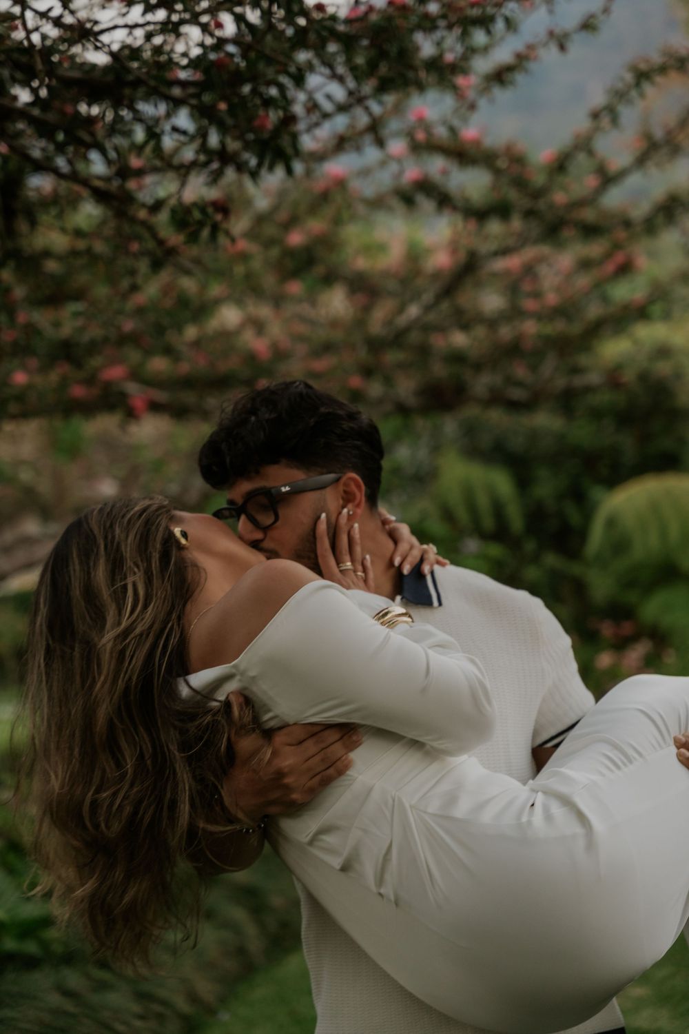 Couple sharing a romantic kiss in a lush garden with pink flowering trees, woman in white outfit being lifted by her partner.