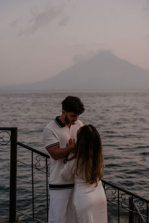 Couple in white embracing on a lakeside railing at dusk with a volcanic mountain silhouette in the background.