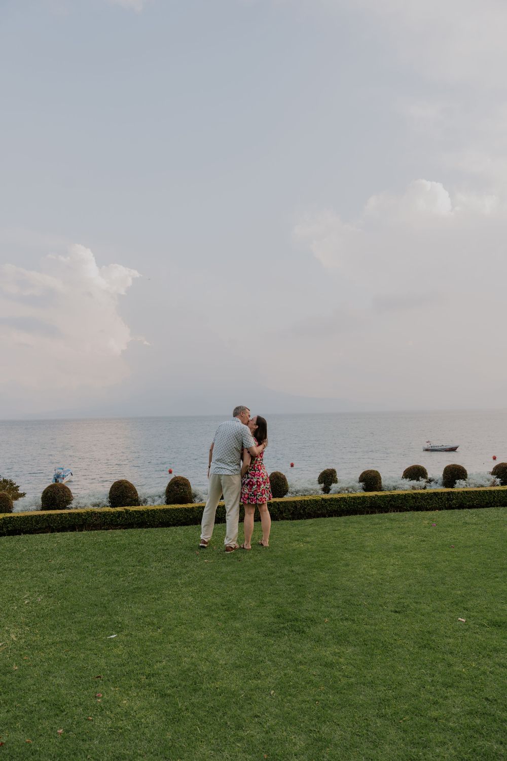 Couple embracing on lush green lawn overlooking calm sea with a boat and manicured hedges under a cloudy sky.
