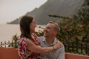 A smiling couple embraces on a scenic terrace with mountain and ocean views in the background.