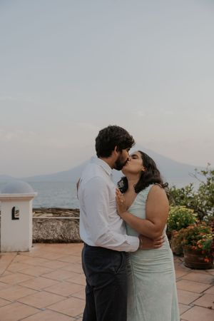 Couple sharing a romantic kiss on a terrace with a mountain and sea view in the background.
