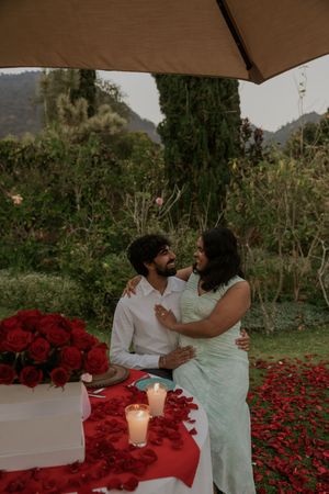 Romantic couple embracing at candlelit outdoor table adorned with red roses and petals in a lush garden setting.