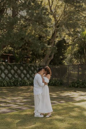 A tender moment between two people in white outfits embracing on a sunlit garden lawn.