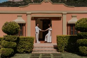 Newlyweds hold hands on the steps of a pink Spanish colonial style building with manicured gardens.
