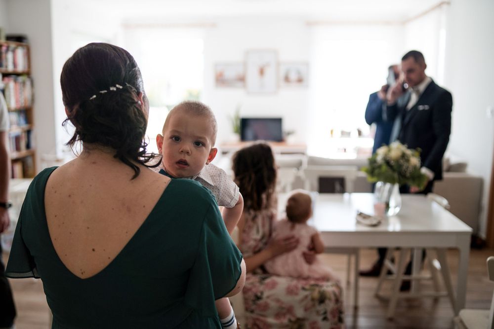 Zöld ruhás nő a kezében egy kisbabával egy fedett esküvői összejövetelen, a háttérben fotóssal.