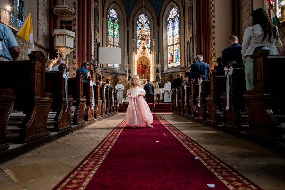 Fiatal virágáruslány rózsaszín ruhában sétál a vörös szőnyeges folyosón egy díszes templomban az esküvői szertartás alatt.