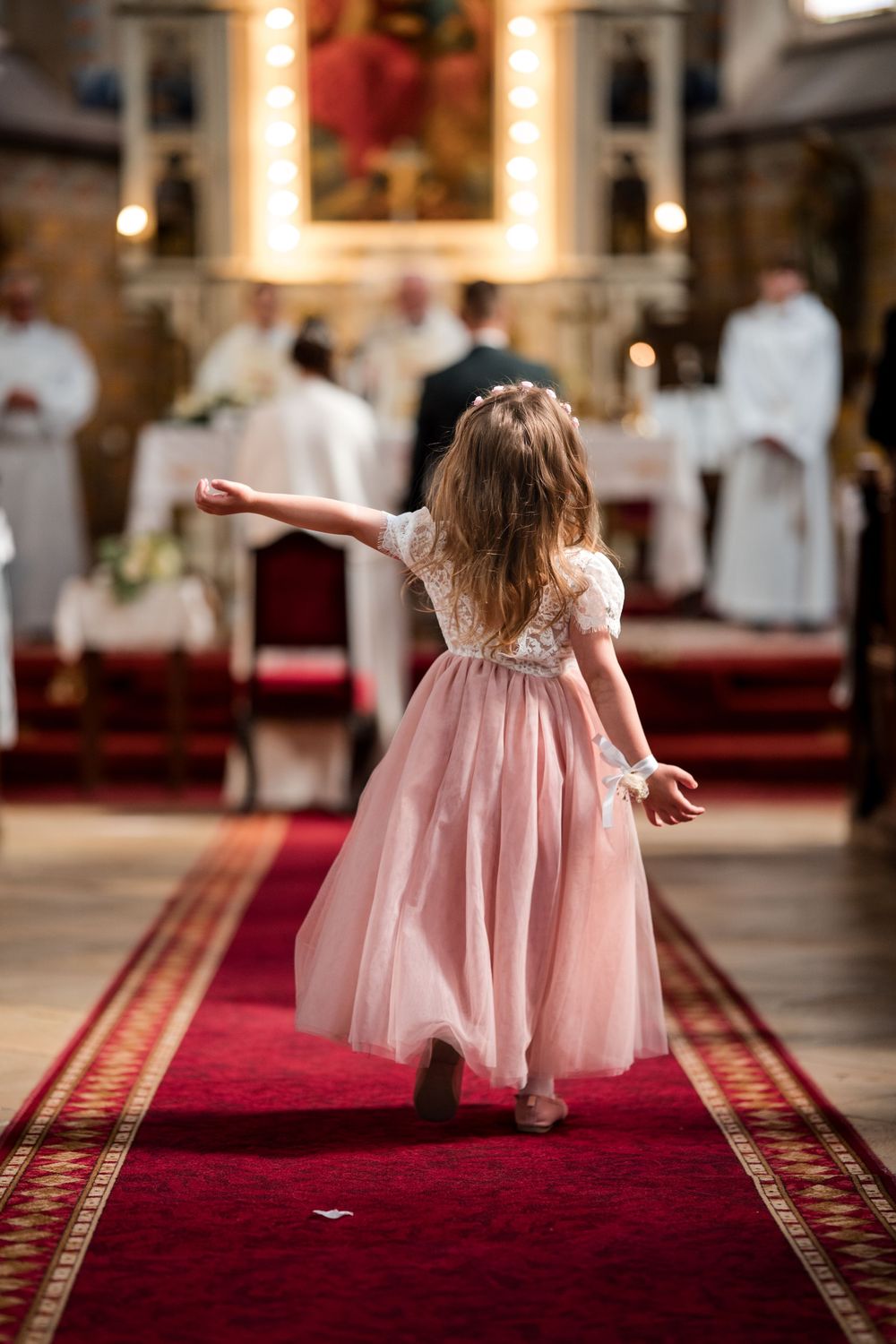 Fiatal virágáruslány rózsaszín tüllruhában sétál végig a vörös szőnyeges folyosón a templomi esküvői szertartáson.