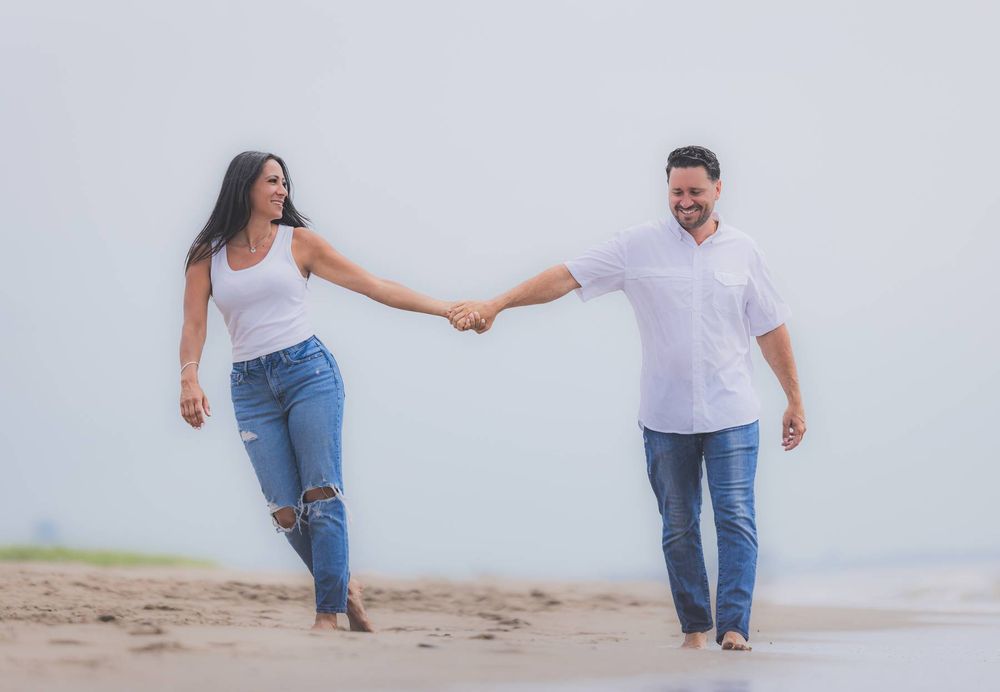 A couple holds hands while walking together on a beach wearing casual outfits of white shirts and jeans.