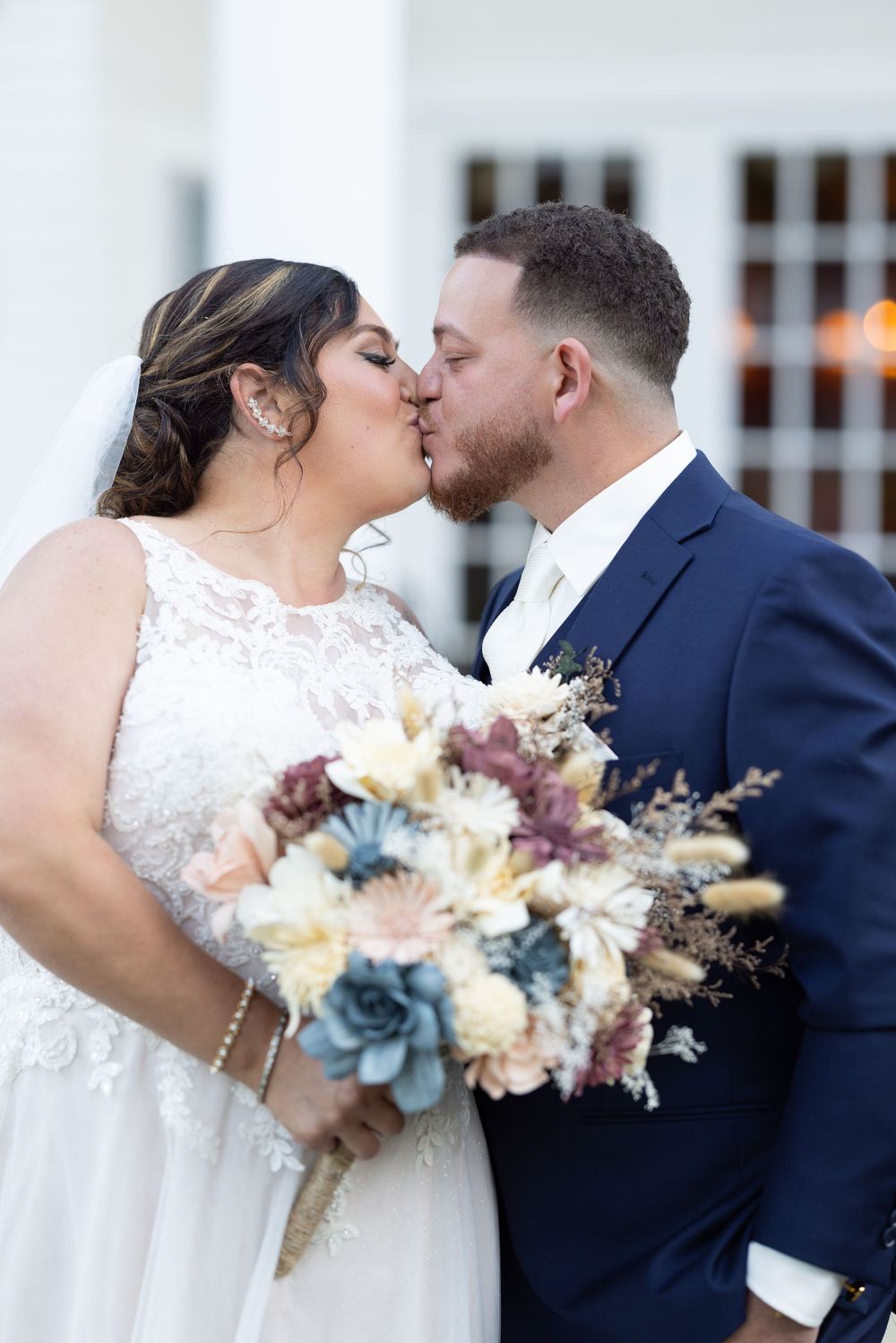 A wedding couple shares a kiss while holding a bouquet of mixed flowers in pastel colors.